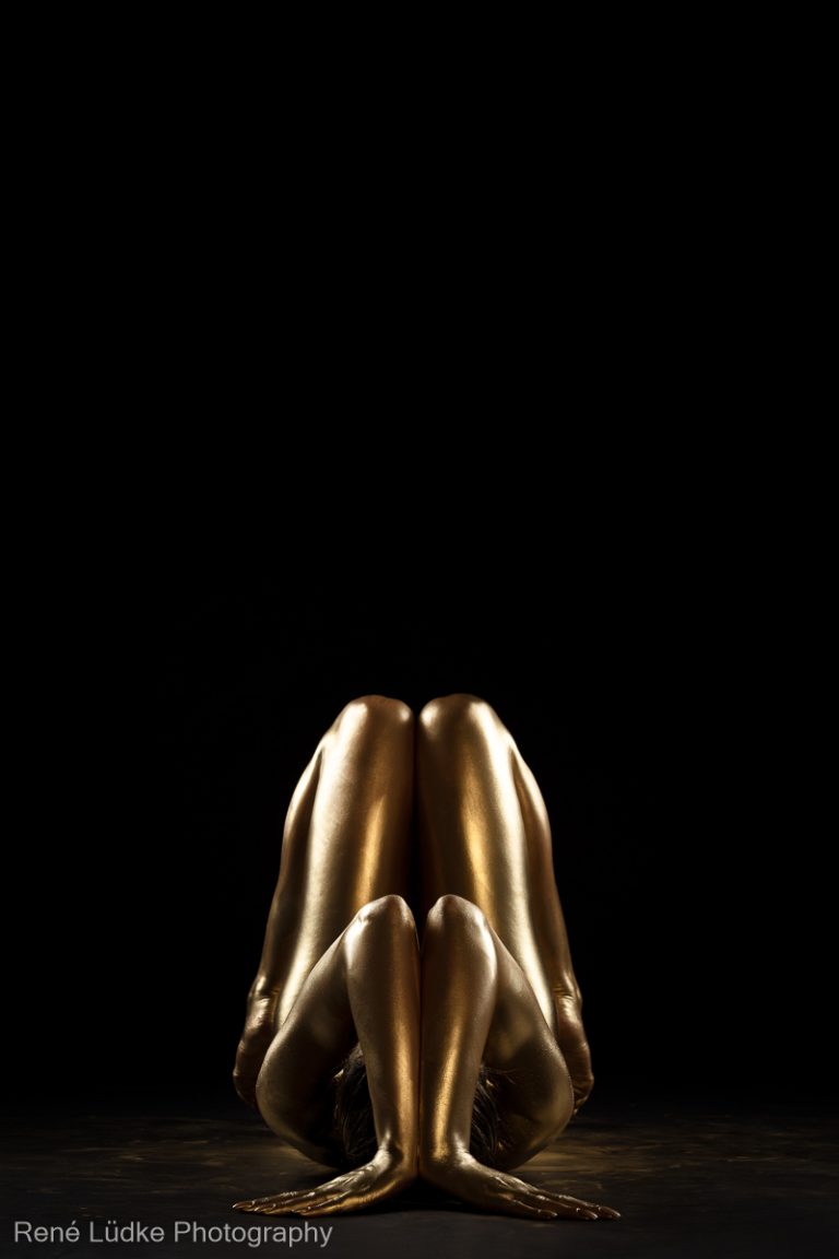 Bodypainting in Gold, auf dem Rücken liegend, Arme und Beine angewinkelt, symmetrische Pose.