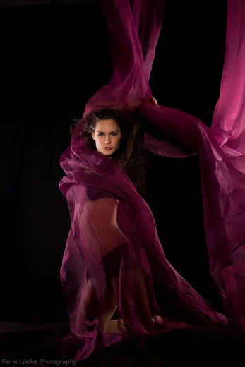 Bild in Farbe von einer Frau in lila Dessous, der Wind umhüllt ihren Körper mit einem halbdurchsichtigen in lila gehaltenen Tuch, sehr dynamisches Bild